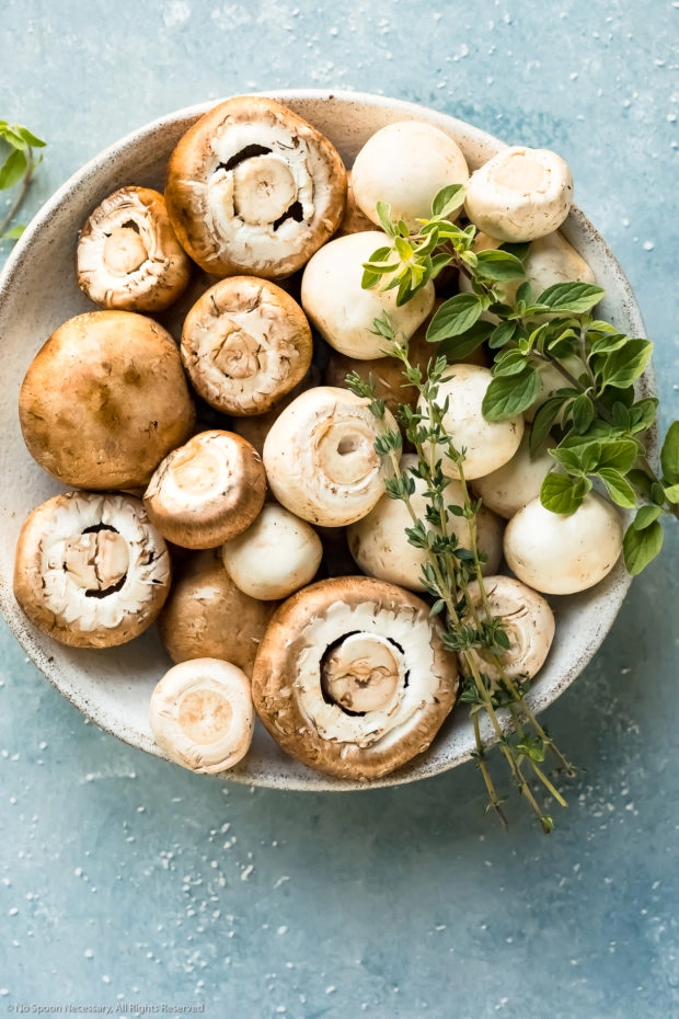 Overhead photo of fresh cremini and button mushrooms, thyme sprigs and oregano sprigs in a white bowl - photo of the main ingredients in the roasted mushroom recipe.