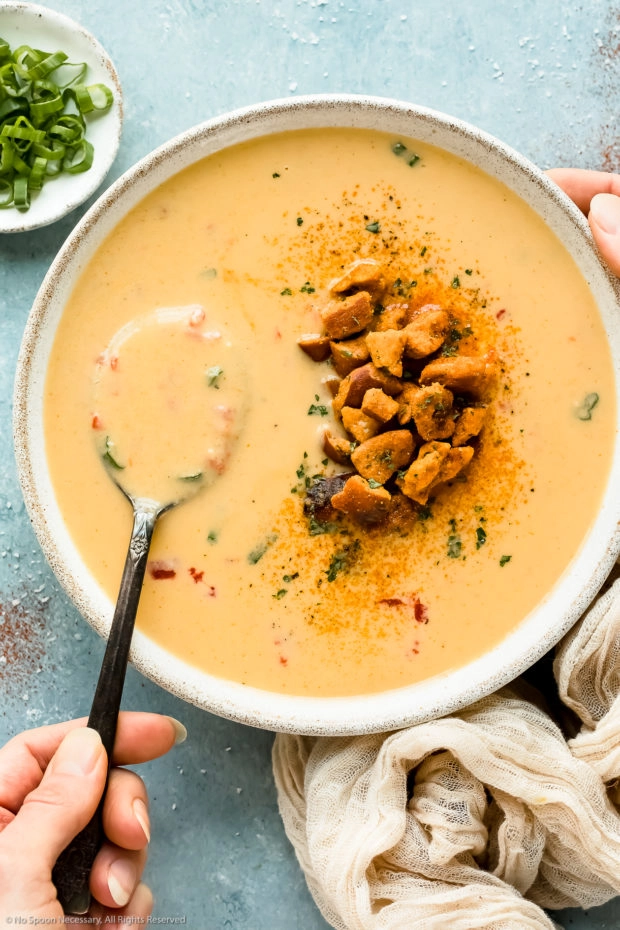 Overhead photo of beer cheddar soup topped with pretzel pieces in a white bowl with a hand holding a spoon inserted into the soup.