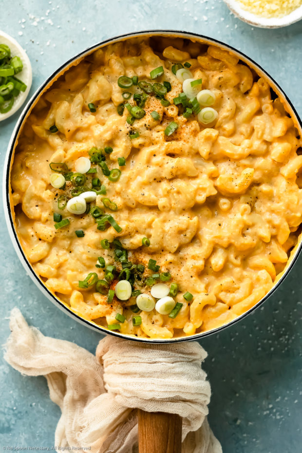Overhead photo of Healthy Stovetop Macaroni and Cheese garnished with sliced scallions in a white saucepan with ramekins of sliced scallions and grated cheese next to the pan.