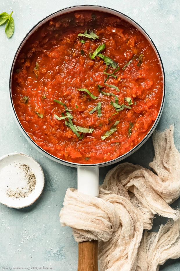 Overhead photo of homemade basil marinara sauce in a white saucepan with a pale tan napkin and a ramekin of salt and pepper next to the pan.