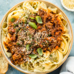 Overhead photo of Slow Cooker Short Rib Ragu Bolognese over papparelle pasta in a neutral colored serving bowl with slices of toasted baguette, fresh basil leaves, a ramekin of grated parmesan cheese and a pale blue linen arranged around the bowl.