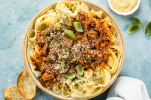 Overhead photo of Slow Cooker Short Rib Ragu Bolognese over papparelle pasta in a neutral colored serving bowl with slices of toasted baguette, fresh basil leaves, a ramekin of grated parmesan cheese and a pale blue linen arranged around the bowl.