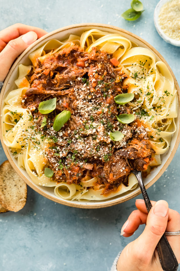 Overhead photo of Beef Bolognese sauce over papparelle pasta in a neutral colored serving bowl with a hand holding the side of the bowl and another hand inserting a fork into the ragu sauce (photo of how to serve ragu bolognese sauce).