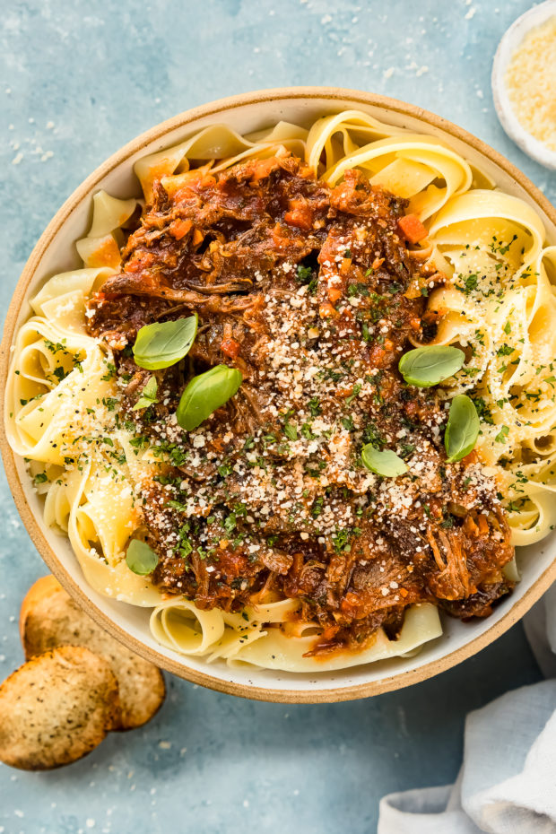 Overhead photo of Slow Cooker Ragu Bolognese over papparelle pasta in a neutral colored serving bowl with slices of toasted baguette, a ramekin of grated parmesan cheese and a pale blue linen arranged around the bowl.