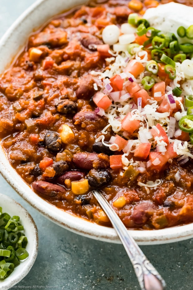 Angled close-up photo of Vegetarian 3 Bean Chili garnished with shredded white cheddar cheese, diced tomatoes and scallions in a white bowl with a spoon inserted into the chili and a ramekin of sliced scallions next to the bowl.