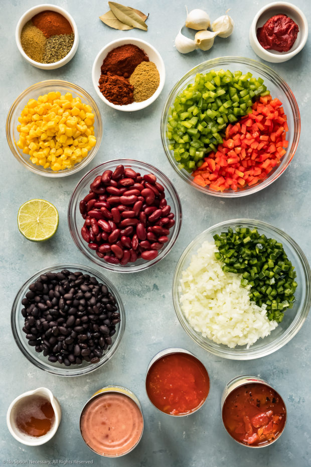 Overhead photo of all the ingredients needed to make three bean chili neatly arranged in individual glass bowls on a blue surface.