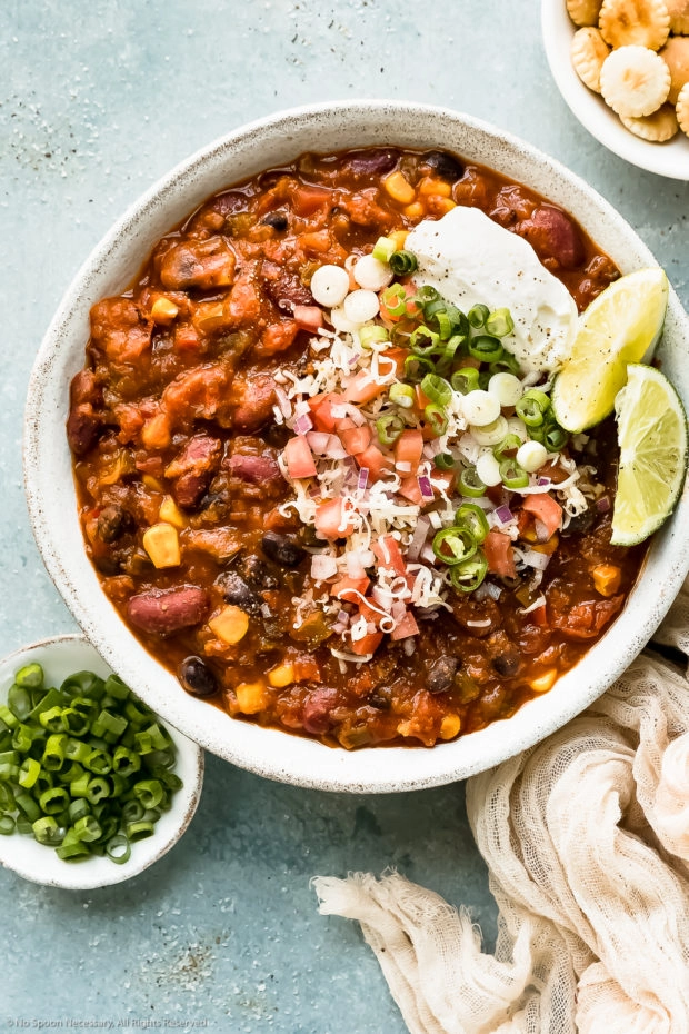 Overhead photo of Vegetarian 3 Bean Chili garnished with shredded cheese, diced tomatoes, scallions, lime wedges and a dollop of sour cream in a white bowl; with ramekins of oyster crackers and sliced scallions next to the bowl.