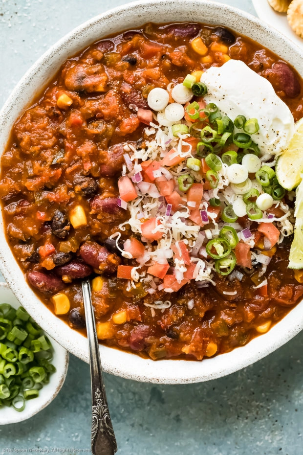 Overhead, up close photo of 3 Bean Chili garnished with shredded cheese, diced tomatoes, sliced scallions and a dollop of sour cream in a white bowl with a ramekin of sliced scallions next to the bowl.