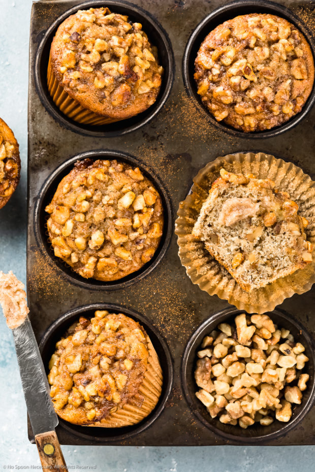 Overhead photo of Banana Nut Muffins in a metal 6 count muffin pan with one of the muffins cut in half showcasing the interior texture of the muffin. 
