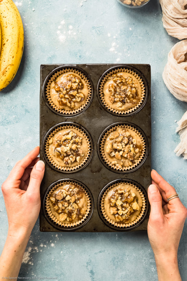 Overhead photo of raw banana nut muffin batter topped with honey coated walnuts spooned into paper lined muffin tins, with two hands holding the sides of the muffin pan.