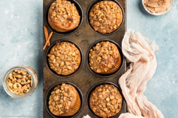 Overhead photo of Banana Nut Muffins in a metal 6 count muffin pan with a ramekin of honey cinnamon butter, jar of walnuts and a light tan linen next to the pan.