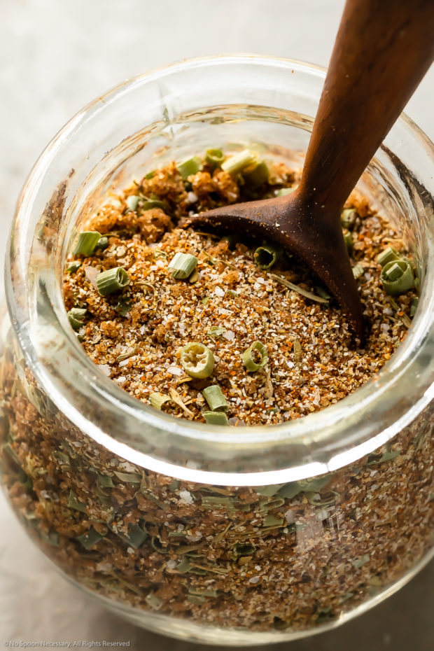 Angled photo of Jamaican Jerk Seasoning in a small glass jar with a wooden spoon inserted into the spice blend.