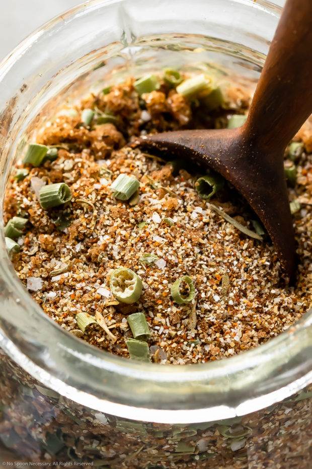 Angled, up-close photo of Homemade Jerk Seasoning in a small glass jar with a wooden spoon inserted into the spice blend.