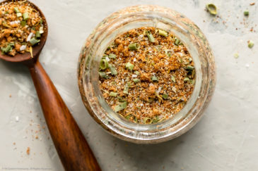 Overhead photo of Jerk Seasoning in a small glass jar with a wooden spoonful of the seasoning mix next to the jar.