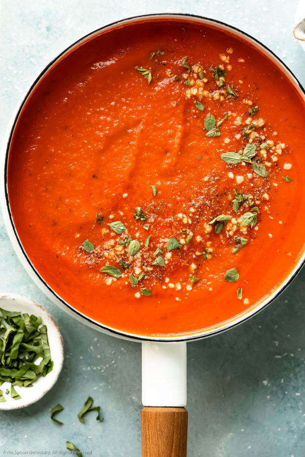 Overhead photo of creamy vodka tomato sauce topped with fresh herbs and grated parmesan in a white saucepan with a ramekin of thinly sliced fresh basil next to the pan.