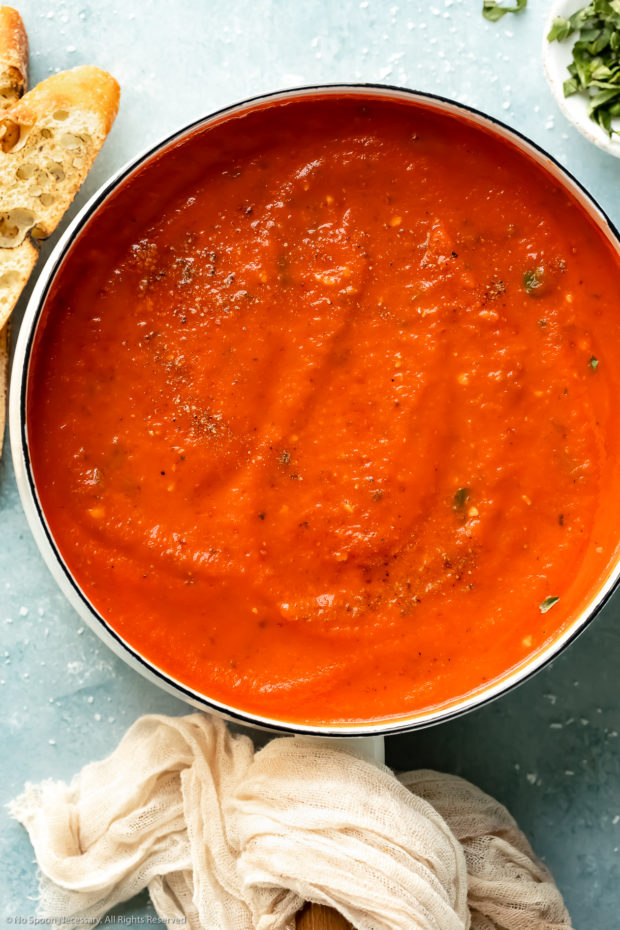 Overhead photo of creamy vodka sauce topped with freshly ground black pepper in a white saucepan with slices of baguette and a ramekin of thinly sliced fresh basil next to the pan.