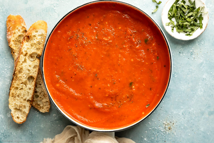 Overhead photo of creamy vodka sauce in a white saucepan with slices of crusty baguette and a ramekin of thinly sliced fresh basil next to the pan.
