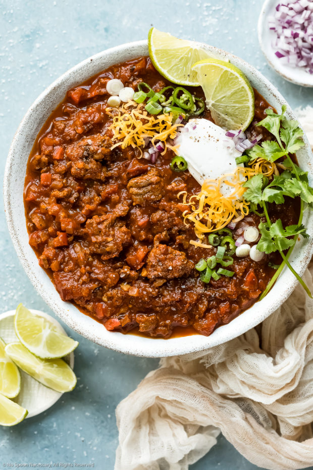 Overhead photo of Texas No Bean Chili garnished with shredded cheese, sour cream, red onion and scallions in a white bowl with a pale neutral colored napkin and ramekins of limes and minced red onions next to the bowl.