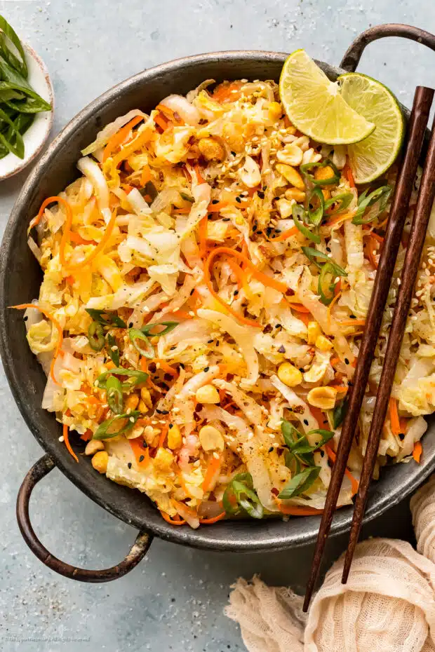 Overhead photo of stir-fried cabbage and carrots garnished with chopped peanuts and lime wedges in an antique skillet with chopsticks resting on the side of the skillet and a ramekin of sliced scallions next to the pan.