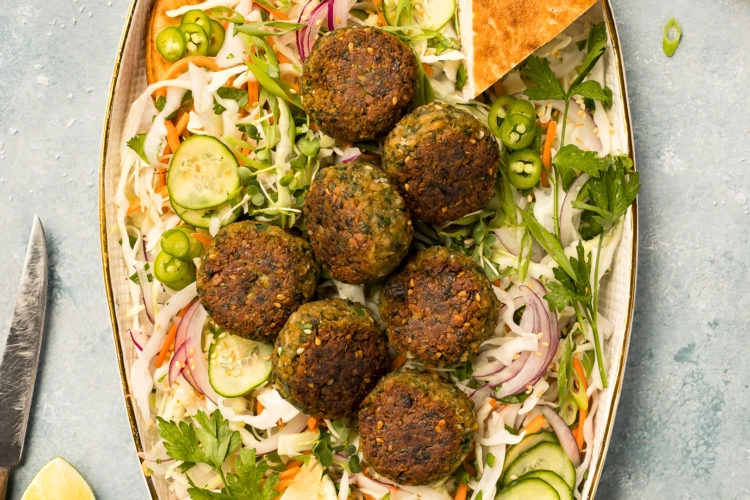 Overhead photo of a platter of Asian Crispy Pan Fried Falafels on a bed of Asian slaw with pita bread; with a pale green linen, knife, lime wedge, ramekin of sliced scallions and small ceramic jar of miso tahini surrounding the platter.