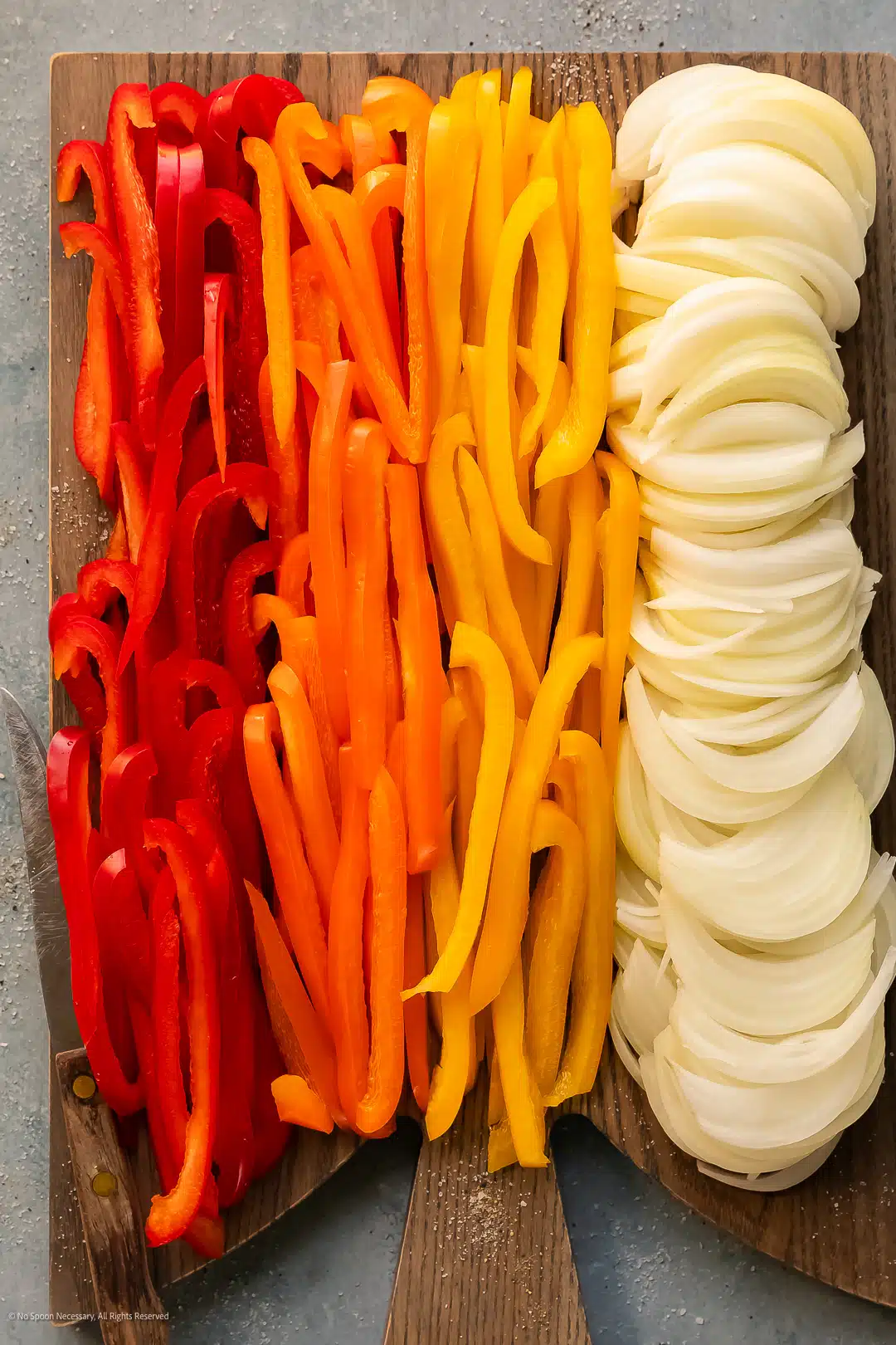 Overhead photo of thinly sliced bell peppers and yellow onions on a clean cutting board.