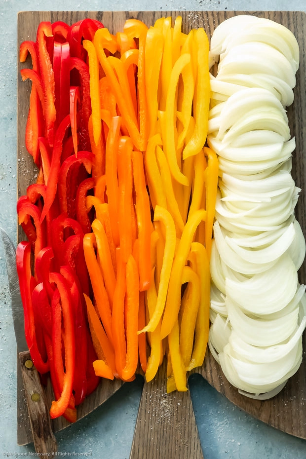 Overhead photo of thinly sliced peppers and onions on a gray wood cutting board - photo of how to cut vegetables for sauteing. 