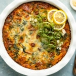 Overhead photo of Crustless Spinach Bacon Quiche garnished with fresh lemon slices and scallions in a white pie pan with a ramekin of sliced scallions and grated parmesan next to the quiche.