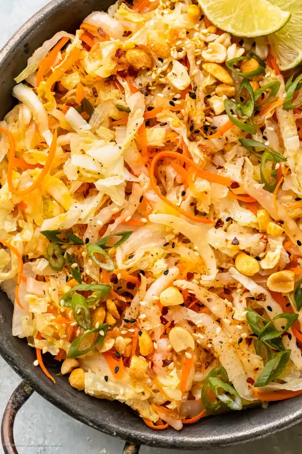 Overhead, up close photo of stir fried cabbage and carrots garnished with chopped peanuts and lime wedges in an antique skillet.