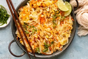 Overhead landscape photo of stir-fried cabbage and carrots garnished with chopped peanuts and lime wedges in an antique skillet with chopsticks resting on the side of the skillet.