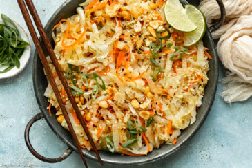 Overhead landscape photo of stir-fried cabbage and carrots garnished with chopped peanuts and lime wedges in an antique skillet with chopsticks resting on the side of the skillet and a ramekin of sliced scallions next to the pan.