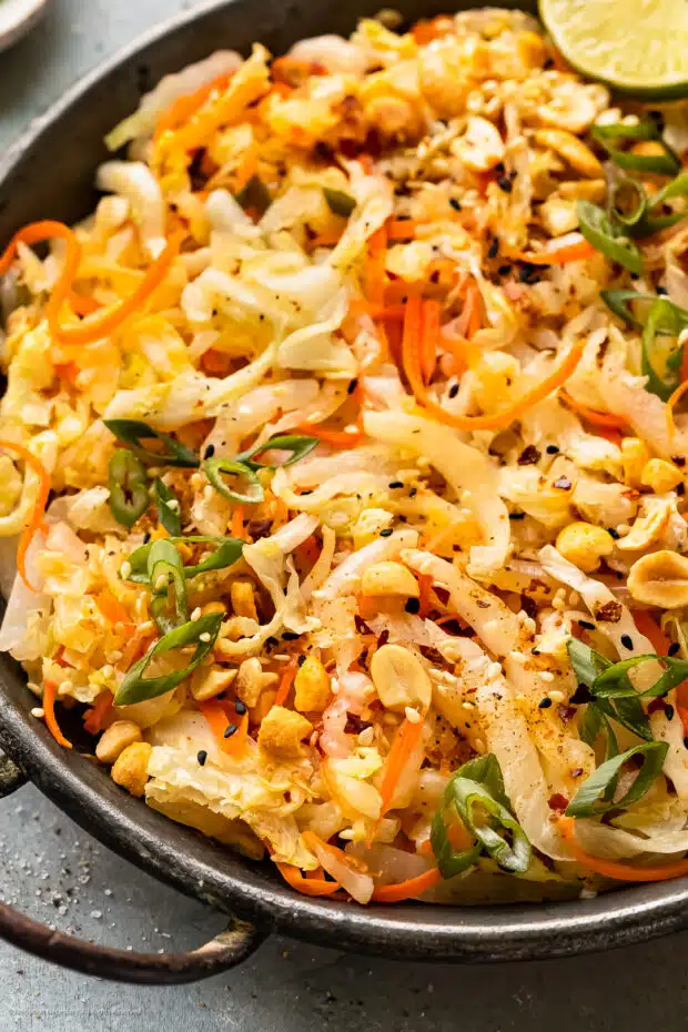 Angled, up close photo of stir fried cabbage garnished with chopped peanuts and lime wedges in an antique skillet.