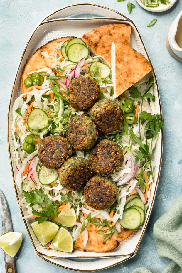 Overhead shot of a platter of Asian Crispy Pan Fried Falafels on a bed of Asian slaw with pita bread; with a knife, lime wedge, ramekin of sliced scallions and small ceramic jar of miso tahini surrounding the platter.