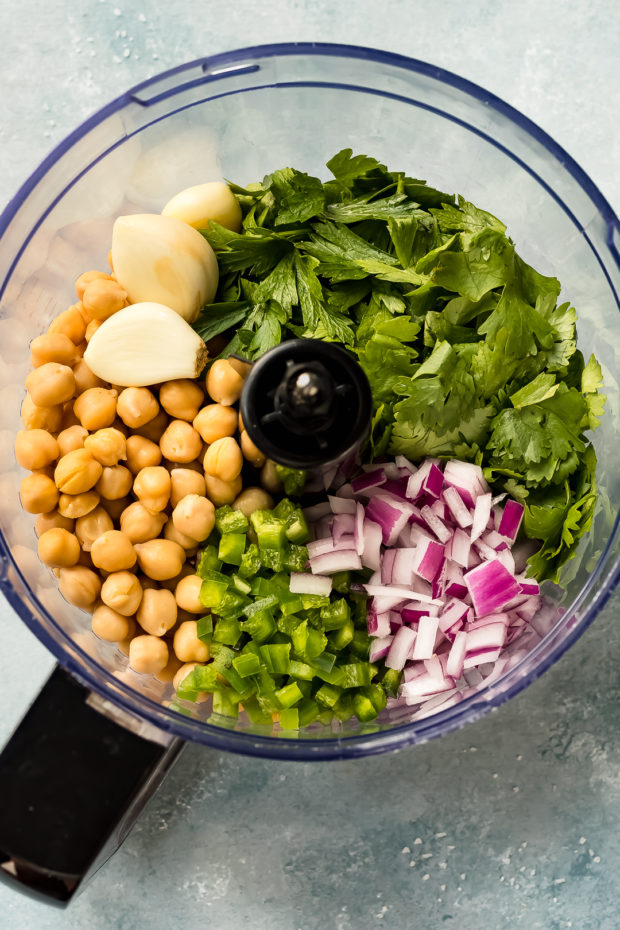 Ingredients to make falafels in a food processor bowl including garlic, chickpeas, onion, cilantro, and other ingredients.