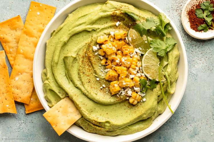 Overhead landscape photo of Avocado Hummus topped with charred corn kernels, crumbled cheese, lime wedges and fresh cilantro in a large white serving bowl with pita strips and a ramekin of sriracha powder next to the bowl.