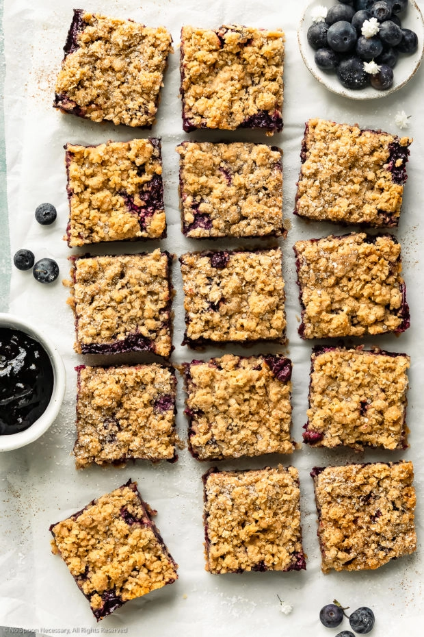 Overhead photo of blueberry crumb bars cut into squares on a large piece of parchment paper with a ramekins of fresh blueberries and homemade blueberry pie filling next to the bars.
