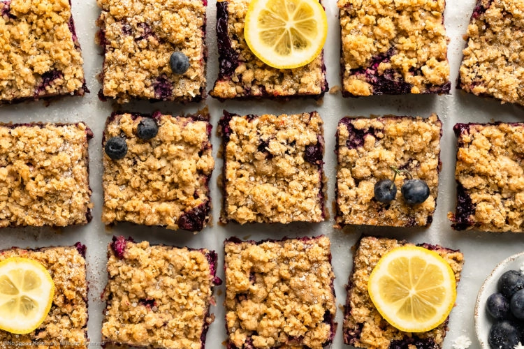 Overhead landscape photo of Lemon Blueberry Crumb Bars cut into squares, sprinkled with powdered sugar and garnished with a couple slices of fresh lemon on a piece of white parchment paper.