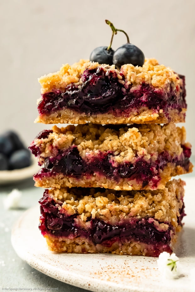 Straight on photo of a stack of three homemade blueberry pie bars on a plate with fresh blueberries garnishing the top.
