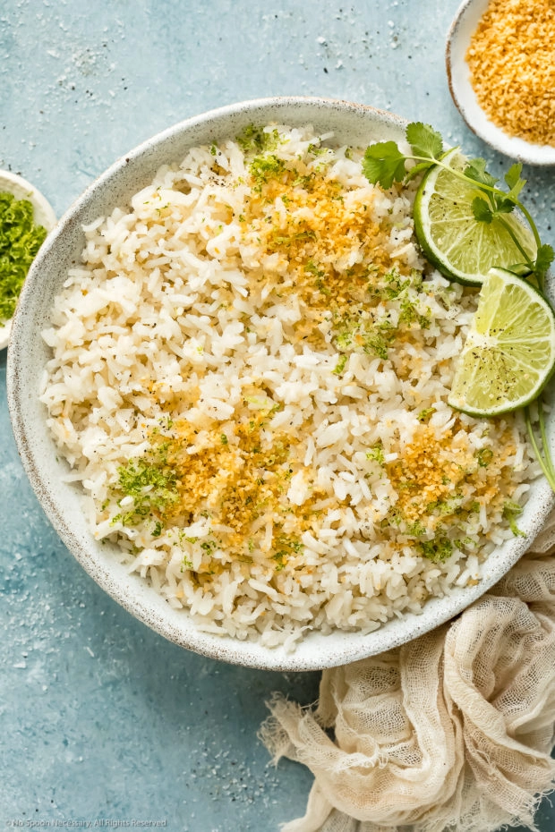 Overhead photo of easy coconut rice garnished with toasted coconut flakes and fresh lime zest in a white bowl with ramekins of lime zest and coconut flakes next to the bowl.