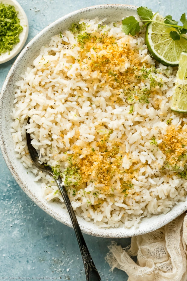 Overhead photo of coconut lime Jasmine rice garnished with fresh lime zest and toasted coconut flakes in a white bowl with a serving spoon inserted into the rice and a ramekin of fresh lime zest next to the bowl.
