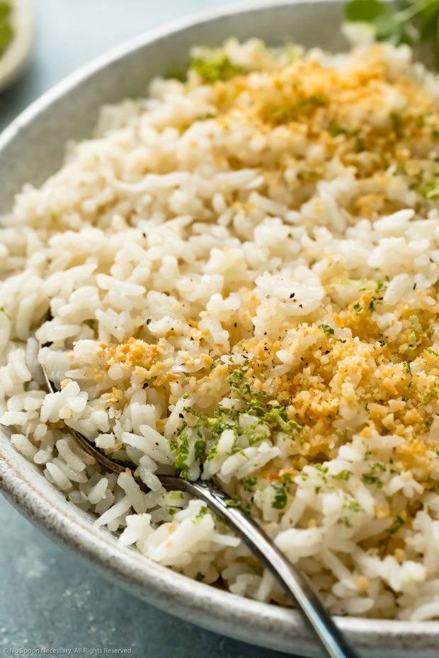 Angled, up-close photo of coconut milk Jasmine rice garnished with fresh lime zest and toasted coconut flakes in a white bowl with a serving spoon inserted into the rice. 