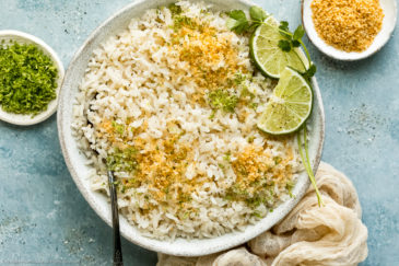 Overhead photo of easy coconut rice garnished with toasted coconut flakes and fresh lime zest in a white bowl with a serving spoon inserted into the rice and ramekins of lime zest and coconut flakes next to the bowl.