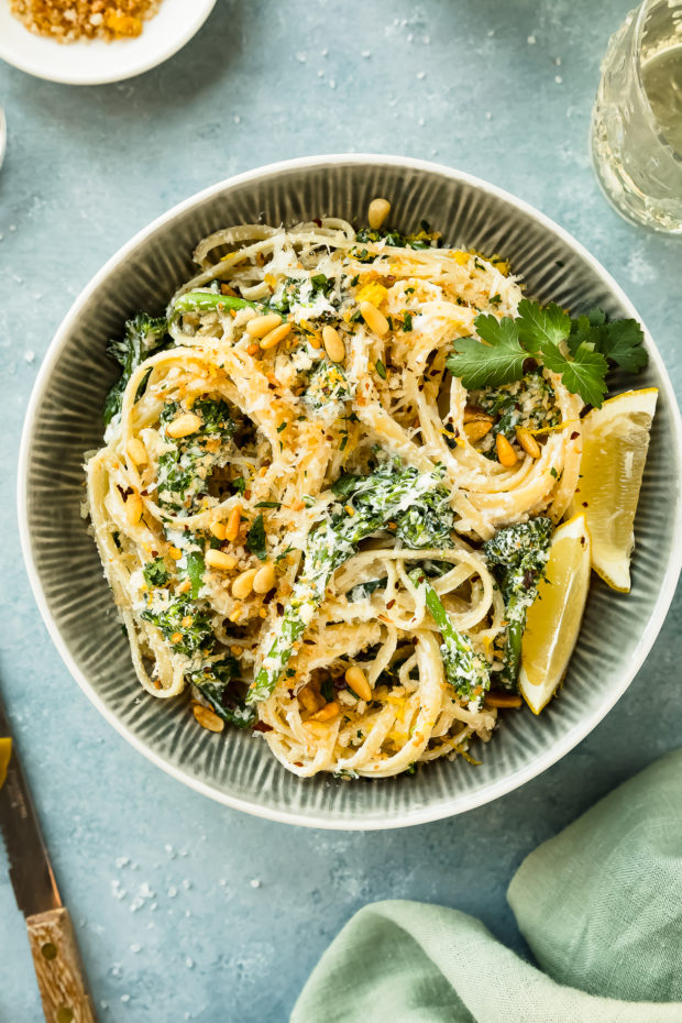 Overhead photo of Creamy Broccoli Pasta in a textured blue bowl with a green napkin, lemon wedges and glass of white wine arranged around the bowl.