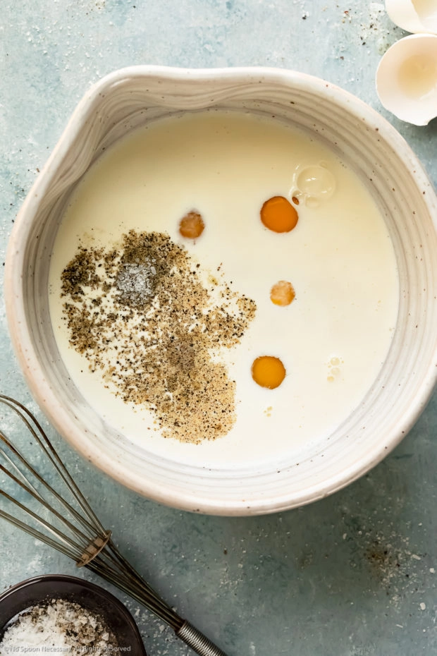 Overhead photo of eggs, half-and-half, salt and pepper in a large white mixing bowl with egg shells and a whisk next to the bowl - photo of preparing egg custard filling for quiche.