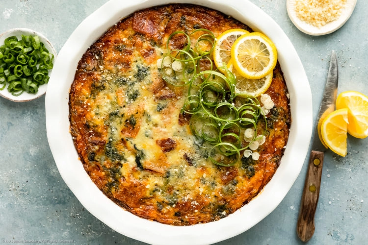 Overhead photo of a Crustless Spinach Quiche garnished with fresh lemon slices and scallions in a white pie pan with a ramekin of sliced scallions and grated parmesan next to the quiche.