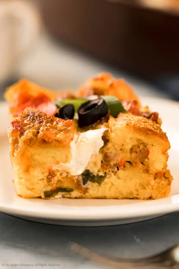 Straight on, up-close shot of the side of a slice of breakfast casserole garnished with olives and scallions with a cup of coffee and a baking dish of the strata blurred in the background.