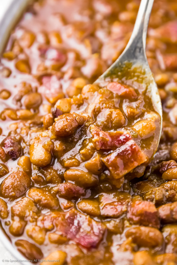 Close-up angled photo of beans and bacon in a galvanized bucket being scooped up with a serving spoon.