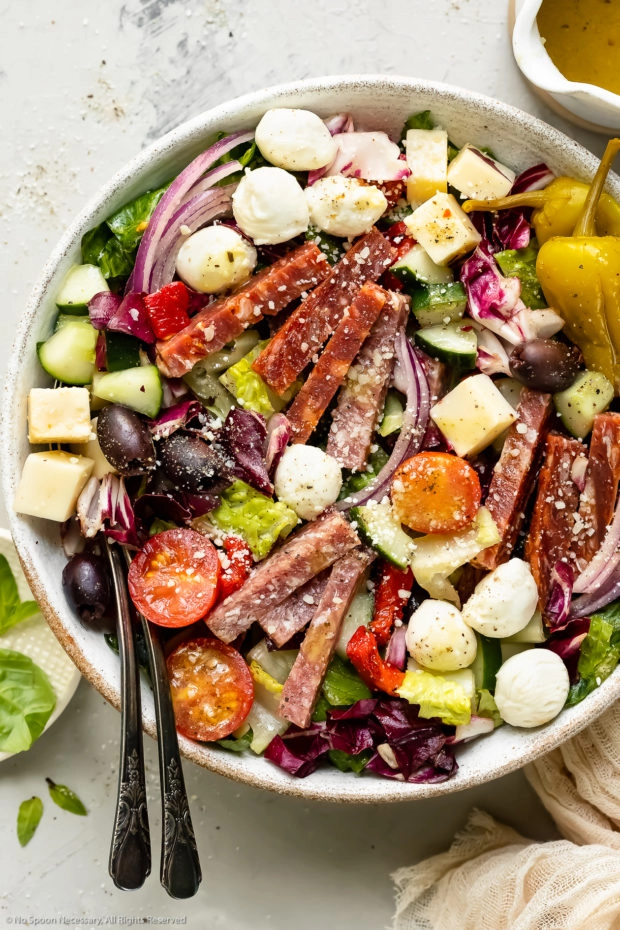 Overhead photo of Italian Chopped Salad in a white bowl with serving spoons inserted into the salad and a jar of homemade Italian dressing and a pale tan napkin arranged around the bowl.