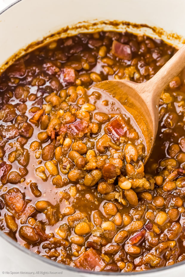 Angled photo of bacon baked beans in a large Dutch oven with a wood serving spoon lifting up the beans. 