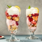 Straight on, landscape shot of a Strawberry Shortcake Sundae with another sundae directly next to it and slightly blurred, and spoons, coarse salt, and a small ramekin of sliced strawberries surrounding the sundae glass.