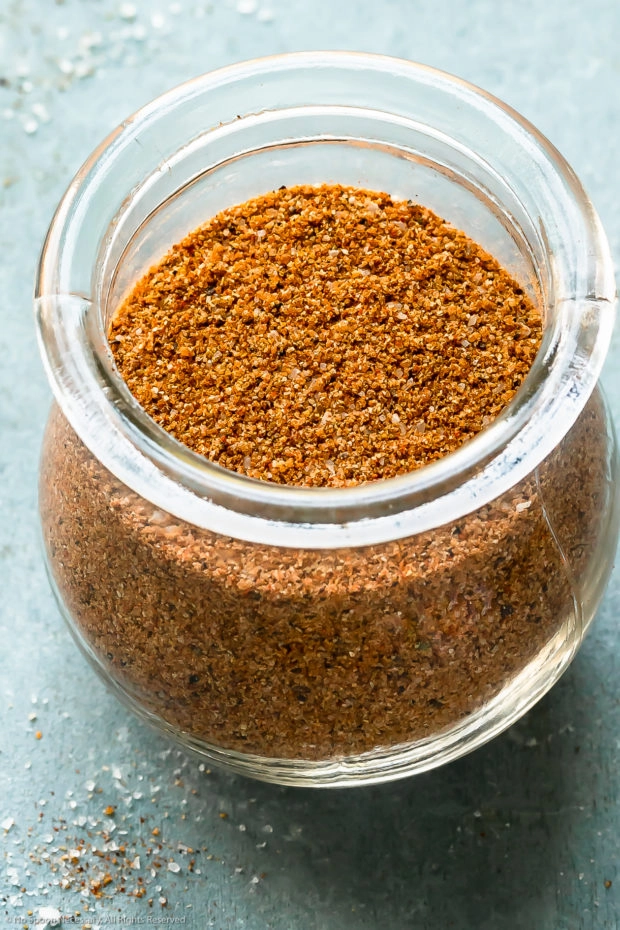 Angled photo of BBQ Dry Rub in a glass jar on a pale blue surface.